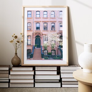 May include: A framed print of a pink brick row house with a dark green door and steps leading up to it. The house has white windows and a tree with white flowers in front of it.