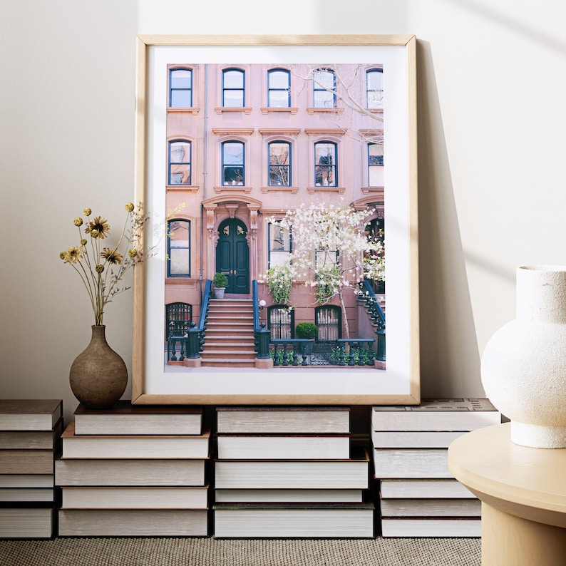 May include: A framed print of a pink brick row house with a dark green door and steps leading up to it. The house has white windows and a tree with white flowers in front of it.