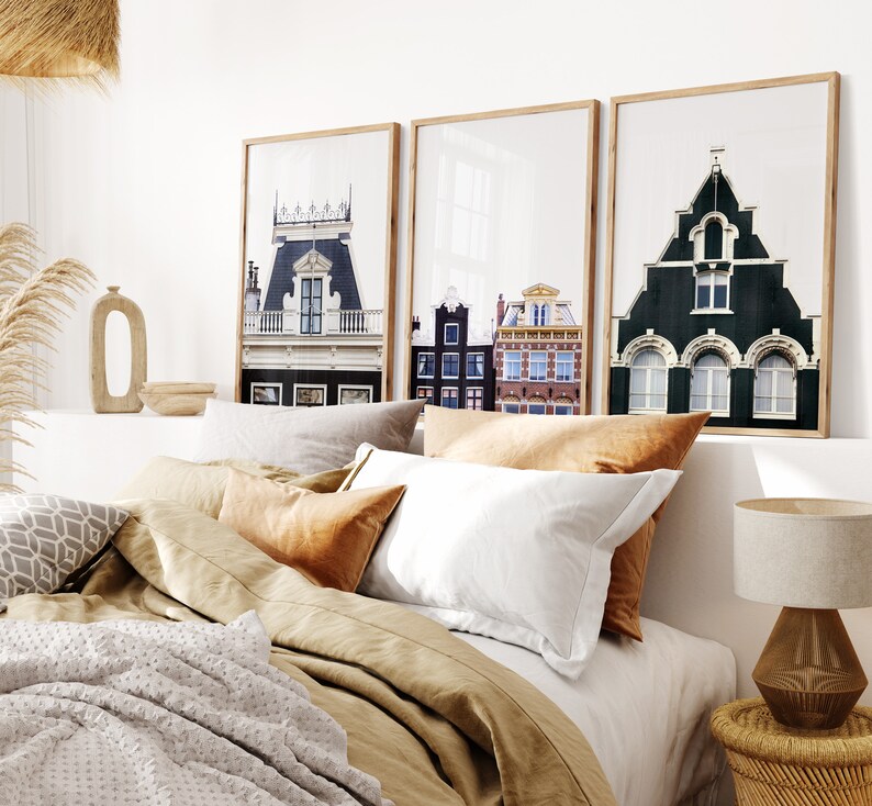 May include: Three framed prints of European cityscapes with colorful buildings and windows. The prints are hanging on a white wall above a bed with tan linen bedding and pillows.