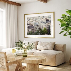 May include: A framed photograph of a Parisian cityscape with a view of buildings and trees. The photo is hanging on a white wall in a living room setting with a beige sofa, a wooden coffee table, and a potted plant.