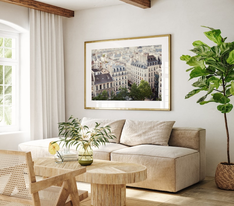 May include: A framed photograph of a Parisian cityscape with a view of buildings and trees. The photo is hanging on a white wall in a living room setting with a beige sofa, a wooden coffee table, and a potted plant.