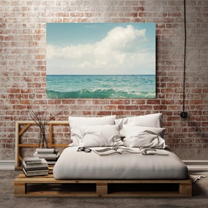 May include: A white bed frame with a wooden pallet base, white bedding, and two white pillows. A stack of books sits on a wooden shelf next to the bed. A framed photograph of a seascape hangs on a brick wall above the bed.