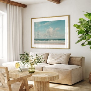 May include: A light beige sectional sofa with two pillows sits in a living room with a large framed photograph of a beach scene with a blue sky and white clouds. A small potted plant with green leaves sits on the floor next to the sofa. A wooden coffee table with a woven design sits in front of the sofa. A wicker chair sits to the left of the sofa.