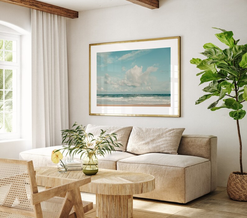 May include: A light beige sectional sofa with two pillows sits in a living room with a large framed photograph of a beach scene with a blue sky and white clouds. A small potted plant with green leaves sits on the floor next to the sofa. A wooden coffee table with a woven design sits in front of the sofa. A wicker chair sits to the left of the sofa.
