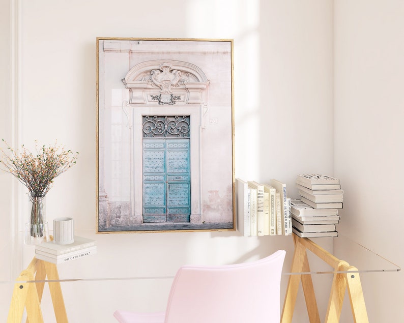 May include: A framed photograph of a vintage, teal blue door with ornate ironwork and a white stone archway. The door is set against a pale pink wall with a faded, textured look.