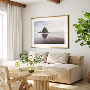 May include: A living room interior with a light beige sectional sofa, a wooden coffee table, a potted ficus tree, and a framed photograph of a rocky island in the ocean.