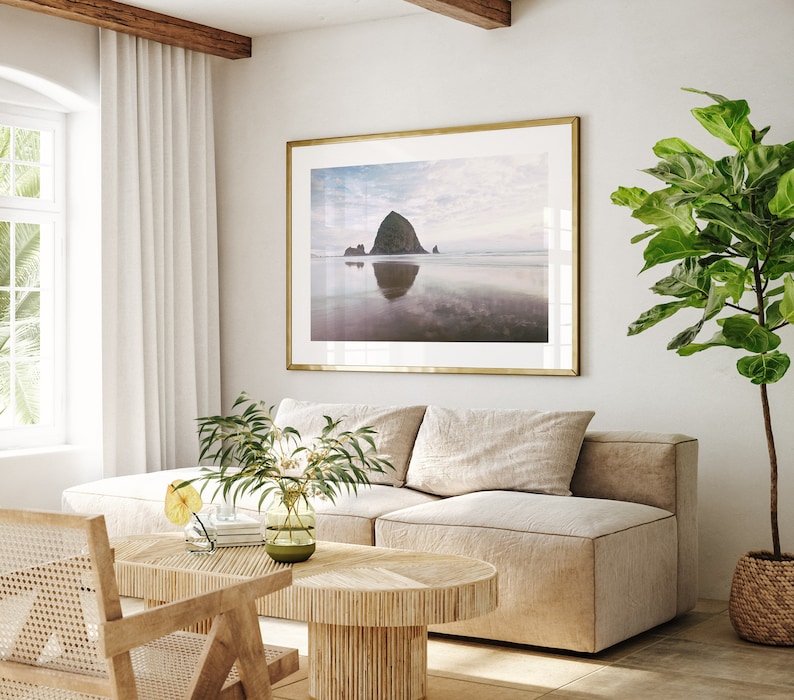May include: A living room interior with a light beige sectional sofa, a wooden coffee table, a potted ficus tree, and a framed photograph of a rocky island in the ocean.