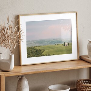 May include: A framed photograph of a landscape with a small white building in the distance. The landscape is green and rolling hills with a few trees. The sky is a soft pink and blue.