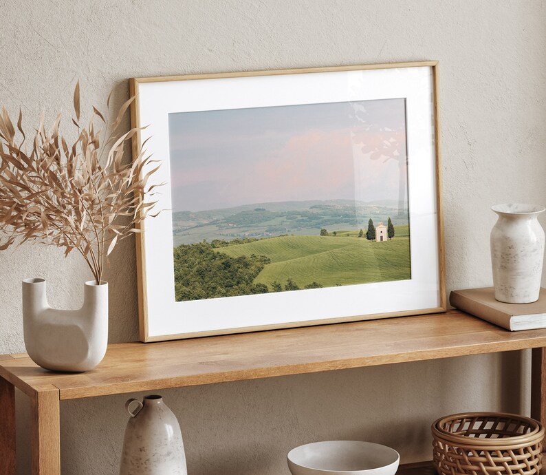 May include: A framed photograph of a landscape with a small white building in the distance. The landscape is green and rolling hills with a few trees. The sky is a soft pink and blue.