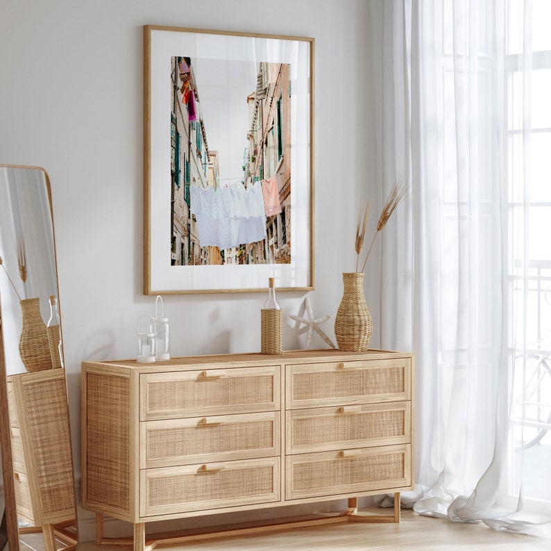 Pu&ograve; includere: Una cassettiera in legno chiaro con sei cassetti, con un fronte in rattan intrecciato. La cassettiera &egrave; decorata con una fotografia incorniciata di una strada stretta in una citt&agrave; europea con biancheria appesa a una corda.