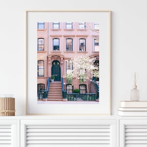 May include: A photograph of a pink brick row house with a black door and white flowers in front. The house has a stoop with steps leading up to the door.