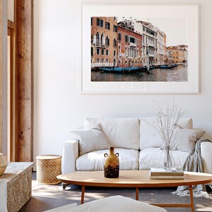 May include: A white couch with a throw blanket and a vase of dried branches sits in front of a framed photograph of a canal in Venice, Italy. The photograph shows colorful buildings lining the canal with gondolas docked along the edge.