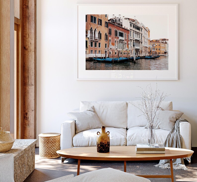 May include: A white couch with a throw blanket and a vase of dried branches sits in front of a framed photograph of a canal in Venice, Italy. The photograph shows colorful buildings lining the canal with gondolas docked along the edge.