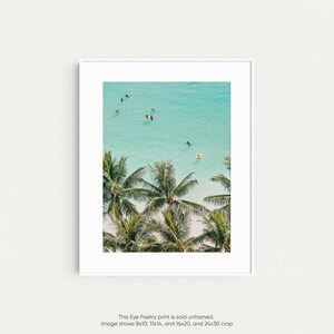 May include: An aerial view of palm trees with lush green leaves against a backdrop of turquoise blue water. People are seen swimming and surfing in the ocean.