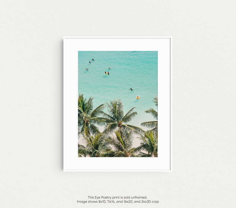 May include: An aerial view of palm trees with lush green leaves against a backdrop of turquoise blue water. People are seen swimming and surfing in the ocean.