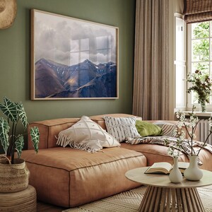 Puede incluir: Un sof&aacute; seccional de cuero marr&oacute;n con almohadas con estampado blanco y negro, una almohada verde y una manta. El sof&aacute; est&aacute; frente a una pared verde con una impresi&oacute;n enmarcada de un paisaje de monta&ntilde;a. Hay flores blancas en un jarr&oacute;n sobre una mesa de caf&eacute; de madera.