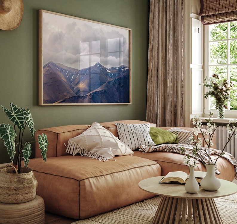 Puede incluir: Un sof&aacute; seccional de cuero marr&oacute;n con almohadas con estampado blanco y negro, una almohada verde y una manta. El sof&aacute; est&aacute; frente a una pared verde con una impresi&oacute;n enmarcada de un paisaje de monta&ntilde;a. Hay flores blancas en un jarr&oacute;n sobre una mesa de caf&eacute; de madera.