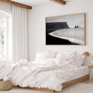 May include: A white bed with a white duvet cover and white pillowcases. The duvet cover has a subtle star pattern. The bed is in a bedroom with a large window and a black and white photograph of a beach scene hanging on the wall.