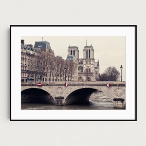 May include: A black and white photograph of the Notre Dame Cathedral in Paris, France. The cathedral is seen from across the Seine River, with a bridge in the foreground. The bridge is made of stone and has several arches. The cathedral is a large, gothic structure with two towers and a spire. The photo is taken on a cloudy day.