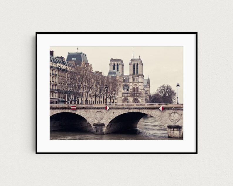 May include: A black and white photograph of the Notre Dame Cathedral in Paris, France. The cathedral is seen from across the Seine River, with a bridge in the foreground. The bridge is made of stone and has several arches. The cathedral is a large, gothic structure with two towers and a spire. The photo is taken on a cloudy day.