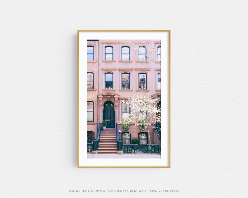 May include: A photograph of a row of brownstone buildings in New York City. The buildings have a pink facade with white trim and black windows. The front door of the building is black and has a set of stairs leading up to it. There is a tree with white blossoms in front of the building.