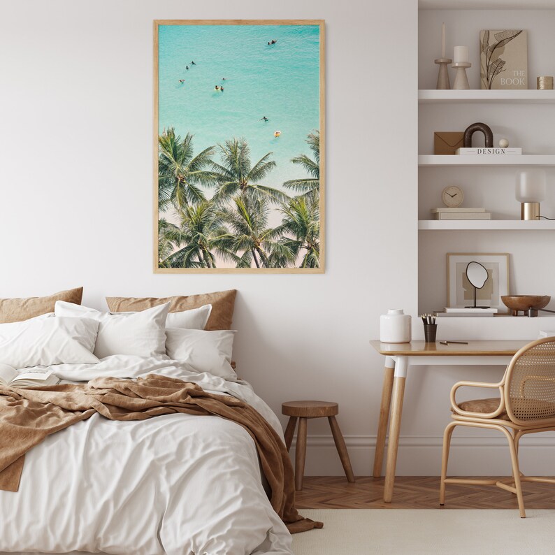May include: A framed photograph of palm trees and turquoise water with people swimming and paddleboarding. The photo is hanging on a white wall in a bedroom setting.