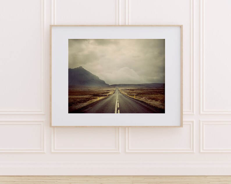May include: A long, straight road stretches into the distance, disappearing into a cloudy sky. The road is surrounded by a vast, open landscape with a mountain in the background.