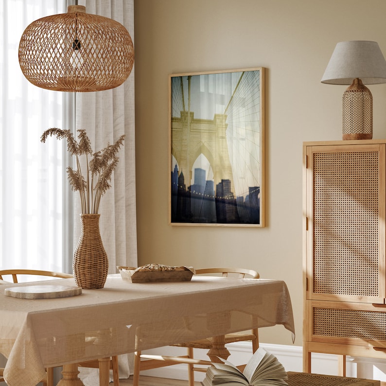 Puede incluir: Una mesa de comedor con un mantel beige, sillas de mimbre y una l&aacute;mpara colgante de mimbre colgando arriba. Una impresi&oacute;n enmarcada de un puente con una ciudad en el fondo cuelga de la pared. Un armario de madera con una puerta de rat&aacute;n tejido est&aacute; en el fondo.
