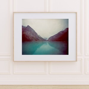May include: A framed photograph of a mountain range reflected in a turquoise lake. The mountains are a dark gray and purple color, and the sky is a light gray. The water is calm and still, reflecting the mountains and sky.