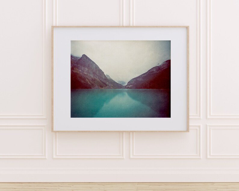 May include: A framed photograph of a mountain range reflected in a turquoise lake. The mountains are a dark gray and purple color, and the sky is a light gray. The water is calm and still, reflecting the mountains and sky.