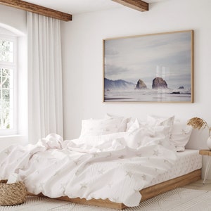 May include: A white bed with a white duvet cover and white pillowcases featuring a subtle star pattern. The bed is made with a wooden frame and is situated in a bedroom with a large window and a framed photograph of a seascape above the headboard.