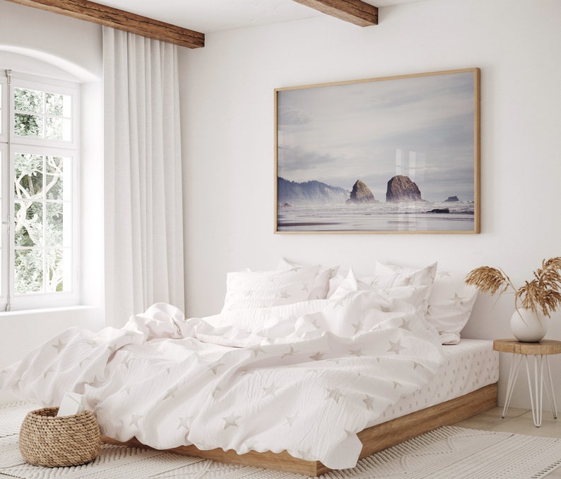 May include: A white bed with a white duvet cover and white pillowcases featuring a subtle star pattern. The bed is made with a wooden frame and is situated in a bedroom with a large window and a framed photograph of a seascape above the headboard.