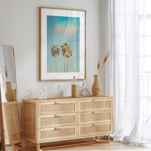 May include: A framed art print featuring three palm trees against a turquoise sky with a rainbow effect. The artwork hangs above a light wood dresser with six woven drawers. Decorative items include a vase, bottle, and starfish.