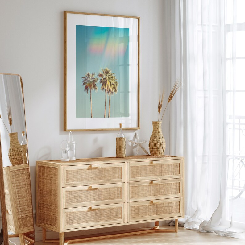 May include: A framed art print featuring three palm trees against a turquoise sky with a rainbow effect. The artwork hangs above a light wood dresser with six woven drawers. Decorative items include a vase, bottle, and starfish.