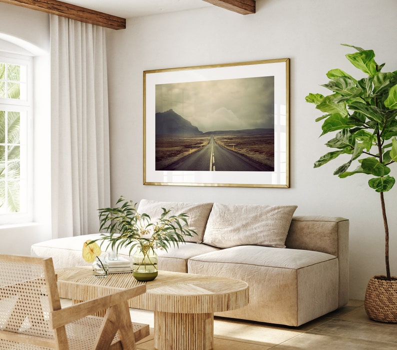 May include: A living room interior with a beige sectional sofa, a wooden coffee table, and a framed photograph of a road leading into a mountain range. A large potted plant is in the corner of the room.