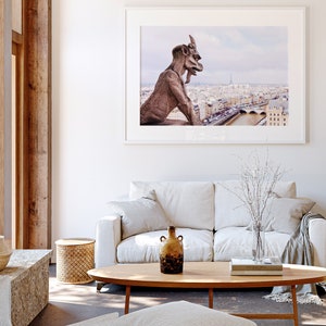 May include: A white sofa with a throw blanket and a vase of white branches sits in front of a framed photograph of a gargoyle overlooking the city of Paris.