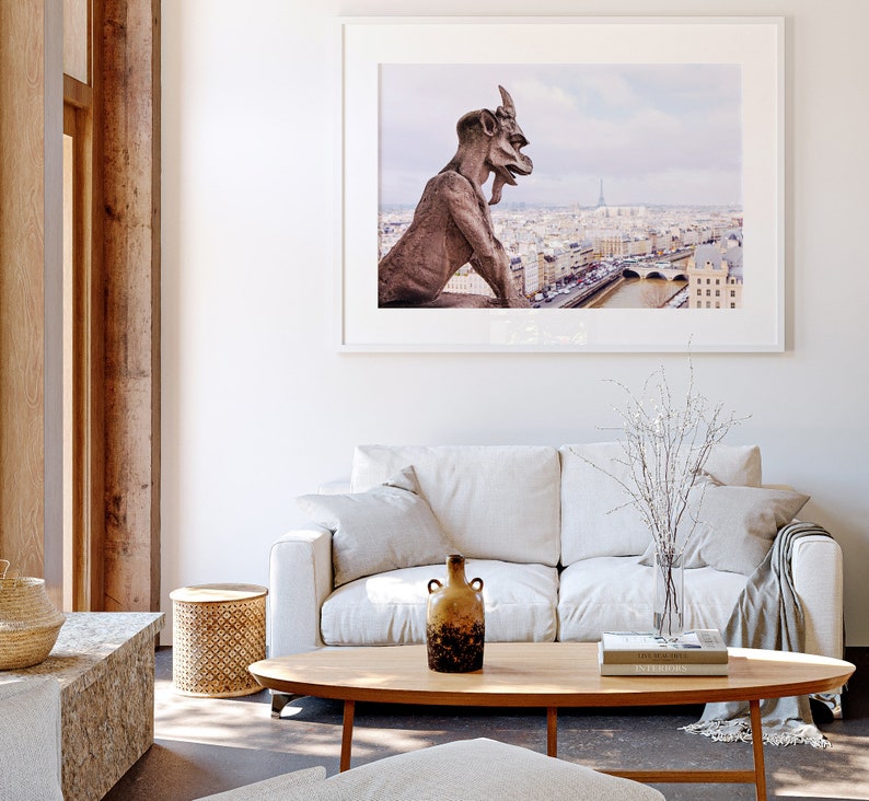 May include: A white sofa with a throw blanket and a vase of white branches sits in front of a framed photograph of a gargoyle overlooking the city of Paris.