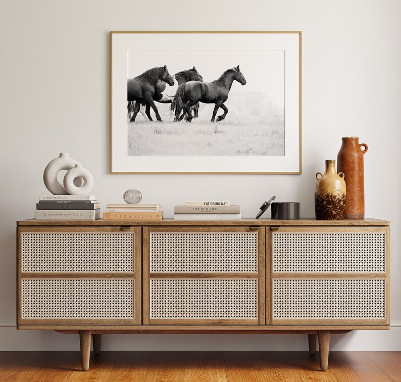 May include: A black and white photograph of a herd of horses running through a field. The photograph is framed in a gold frame and is displayed on a wooden shelf with books and decorative objects.
