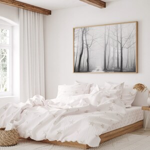 May include: A white bed with a white and gray star patterned duvet cover and matching pillowcases. The bed is made up with a white blanket and has a wooden frame. A black and white photograph of a foggy forest hangs on the wall above the bed.