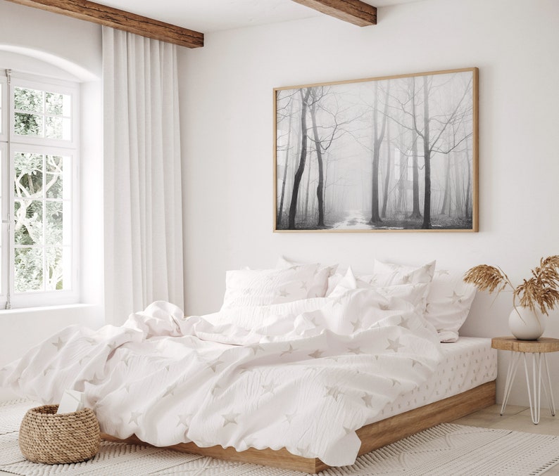 May include: A white bed with a white and gray star patterned duvet cover and matching pillowcases. The bed is made up with a white blanket and has a wooden frame. A black and white photograph of a foggy forest hangs on the wall above the bed.
