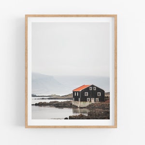 May include: A black wooden house with a red roof sits on a rocky shore by a body of water. The house has white windows and a door. The background is a misty, gray landscape.