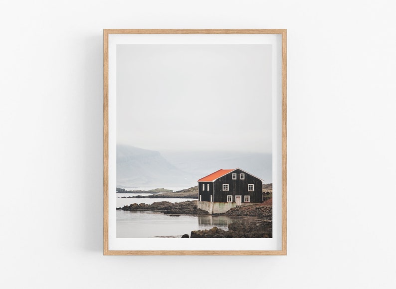May include: A black wooden house with a red roof sits on a rocky shore by a body of water. The house has white windows and a door. The background is a misty, gray landscape.