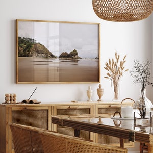 May include: A framed photograph of a beach scene with a rocky coastline and calm water. The photo is hanging on a white wall above a wooden cabinet with woven rattan doors. There are decorative items on the cabinet, including a vase with dried flowers and a glass vase with a plant.