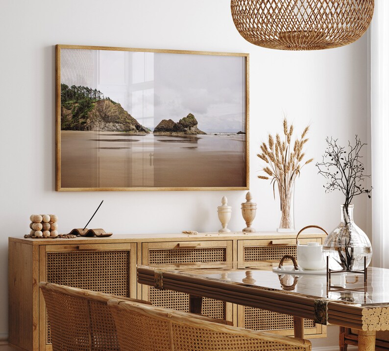 May include: A framed photograph of a beach scene with a rocky coastline and calm water. The photo is hanging on a white wall above a wooden cabinet with woven rattan doors. There are decorative items on the cabinet, including a vase with dried flowers and a glass vase with a plant.