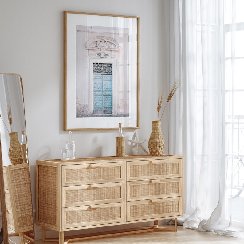 May include: A light wood dresser with six drawers, a framed print of a blue door, a vase with dried flowers, and two glass bottles on top. The dresser is in a room with white walls and white curtains.