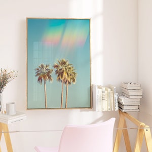 May include: A framed art print featuring two palm trees under a blue sky with a rainbow effect. The artwork has a light wood frame and is displayed in a bright, modern interior setting. The print evokes a tropical, summery vibe.
