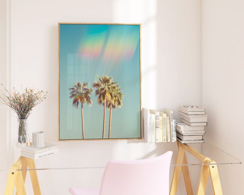 May include: A framed art print featuring two palm trees under a blue sky with a rainbow effect. The artwork has a light wood frame and is displayed in a bright, modern interior setting. The print evokes a tropical, summery vibe.