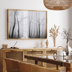May include: A black and white photograph of a foggy forest hangs on a white wall above a wooden cabinet with woven rattan doors. The cabinet is topped with a glass vase filled with dried wheat stalks, a glass vase with a dried branch, and two decorative ceramic vases.