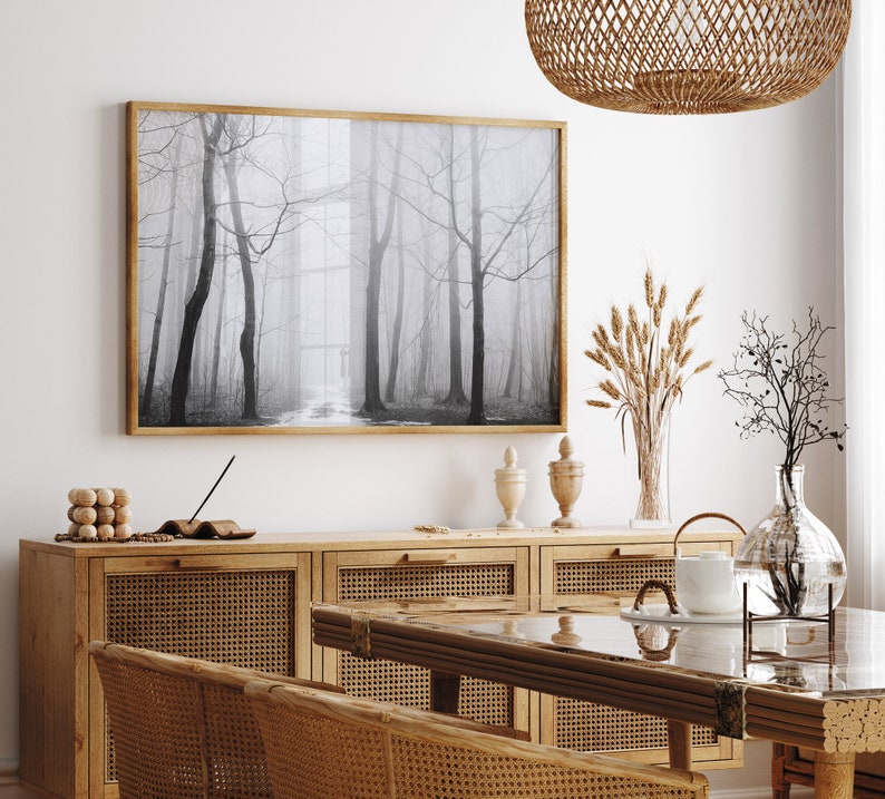 May include: A black and white photograph of a foggy forest hangs on a white wall above a wooden cabinet with woven rattan doors. The cabinet is topped with a glass vase filled with dried wheat stalks, a glass vase with a dried branch, and two decorative ceramic vases.