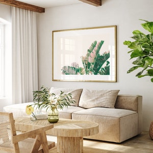 K&ouml;nnte beinhalten: Ein Wohnzimmer-Interieur mit einem beigen Sofa, einem Holz-Couchtisch und einem gerahmten Druck von tropischen Bl&auml;ttern. Der Druck ist in einem goldenen Rahmen und h&auml;ngt an einer wei&szlig;en Wand. In der Ecke des Raumes steht eine gro&szlig;e Topfpflanze.